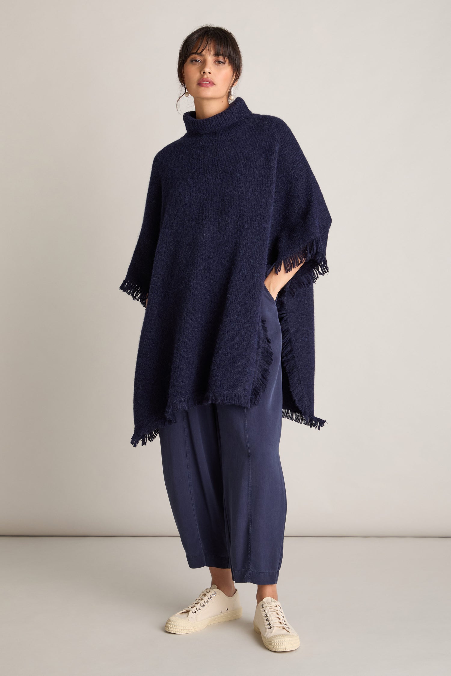 A person stands indoors against a plain background, wearing a luxurious dark blue Cashmere and Alpaca Poncho with fringed edges, paired with blue pants and off-white sneakers.