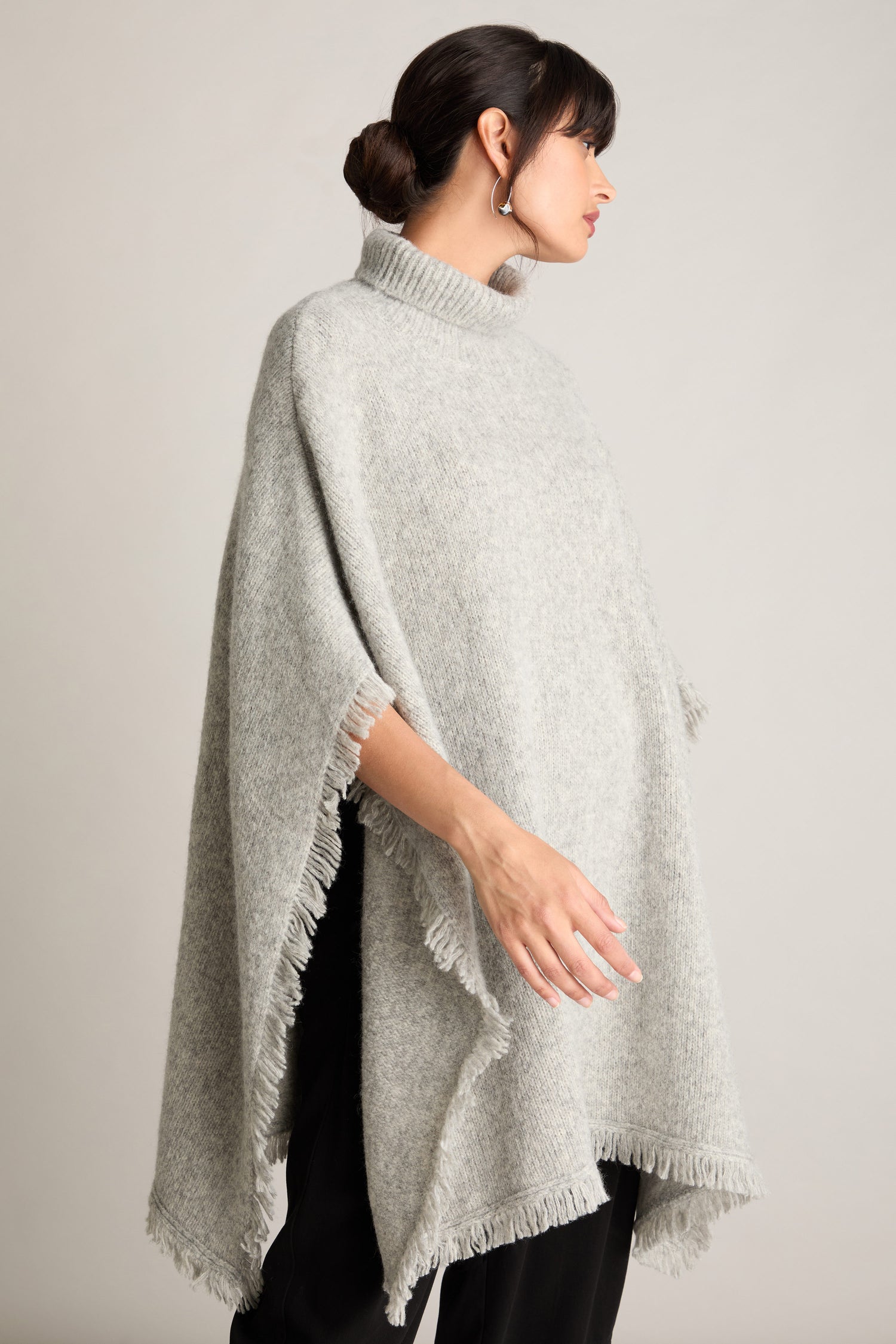 A woman with dark hair pulled back stands in side profile, looking to the left. She is wearing the Cashmere And Alpaca Poncho, a luxurious and comfortable light gray fringed piece, over black pants.