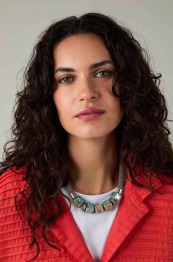 A woman with curly dark hair wears a red textured jacket, a white top, and the Short Marble Cube Necklace in taupe and teal, looking directly at the camera against a neutral background.