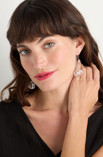A woman with wavy brown hair and a black top touches her neck and looks at the camera, showcasing Silver Scribble Earrings.