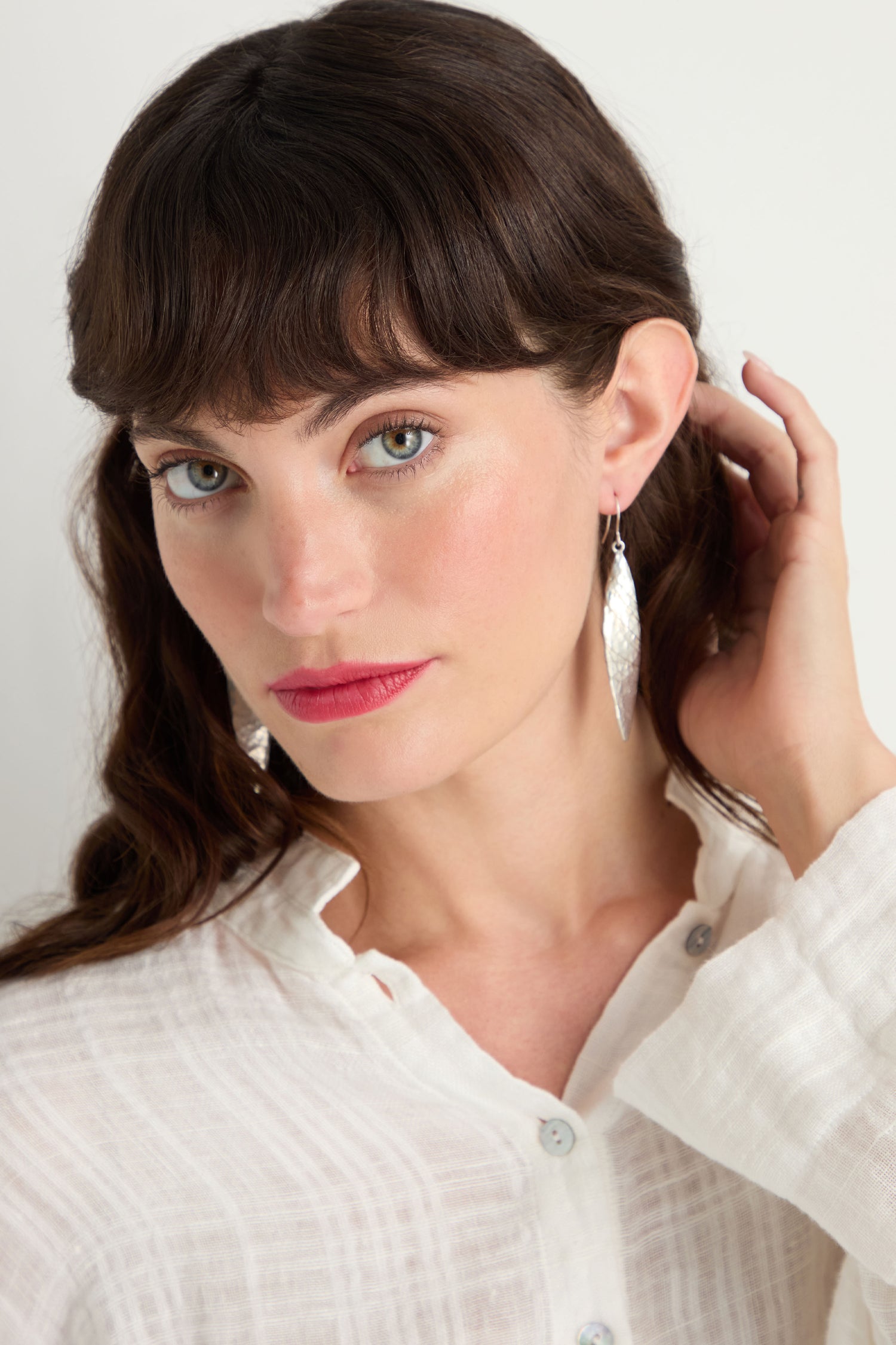 A woman with long brown hair and bangs wears a white button-up shirt and Hammered Silver Leaf Earrings, looking at the camera while touching her hair.