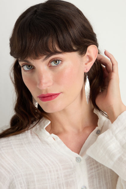 A woman with long brown hair and bangs wears a white button-up shirt and Hammered Silver Leaf Earrings, looking at the camera while touching her hair.