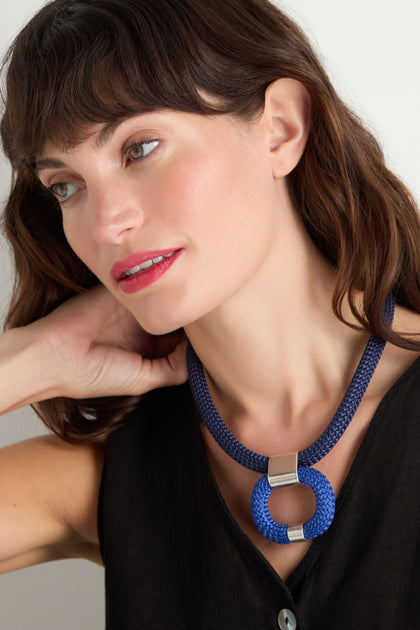 Woman with wavy brown hair wearing a sleeveless black top and the Navy Cord Hoop Necklace, looking to the side with one hand touching her neck.