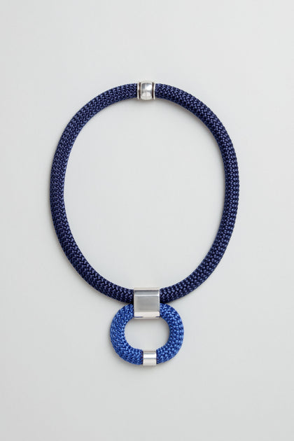 The Navy Cord Hoop Necklace features a navy cord, silver clasp, and a blue and silver circular hoop pendant at the center. Displayed on a light gray background, this piece adds bold elegance to any outfit.