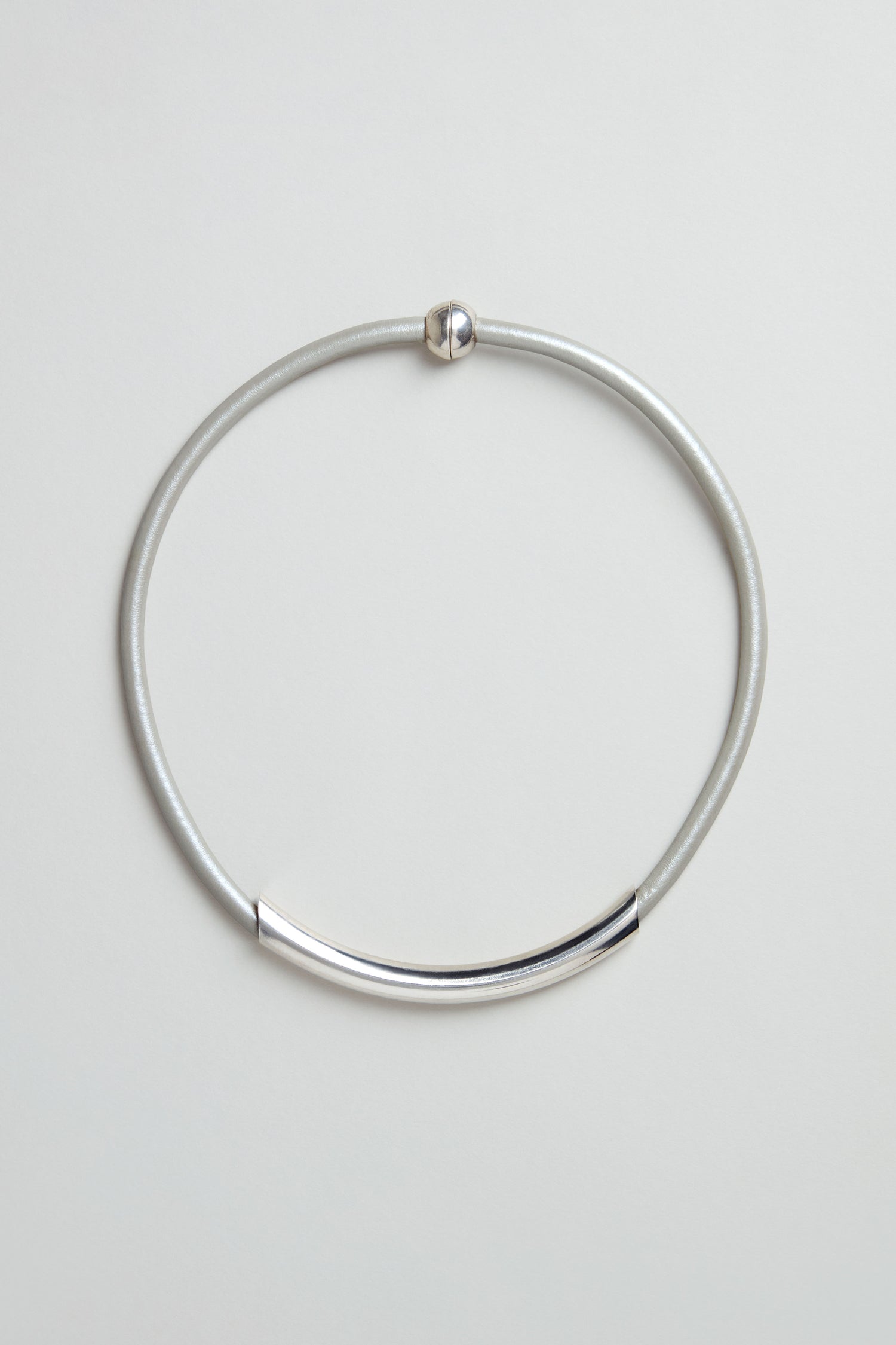 The Grey Leather Cord Necklace is a simple, minimalist piece featuring a curved metal bar and round clasp, presented on a plain light background.