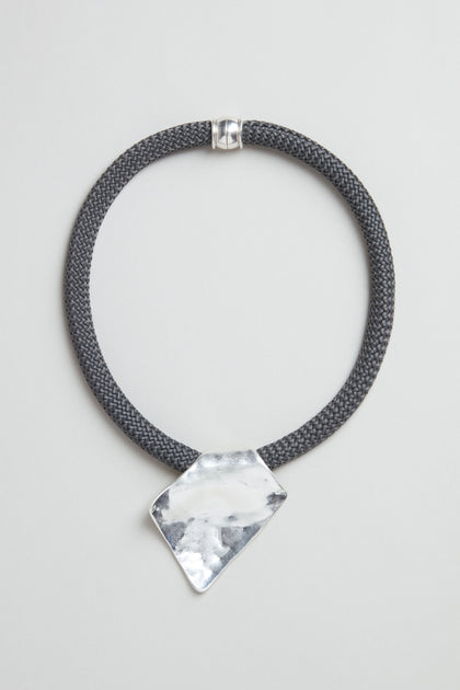 The Hammered Silver Pendant Cord Necklace features a black braided cord, an irregularly-shaped polished hammered silver pendant, and a silver clasp, shown on a light gray background.