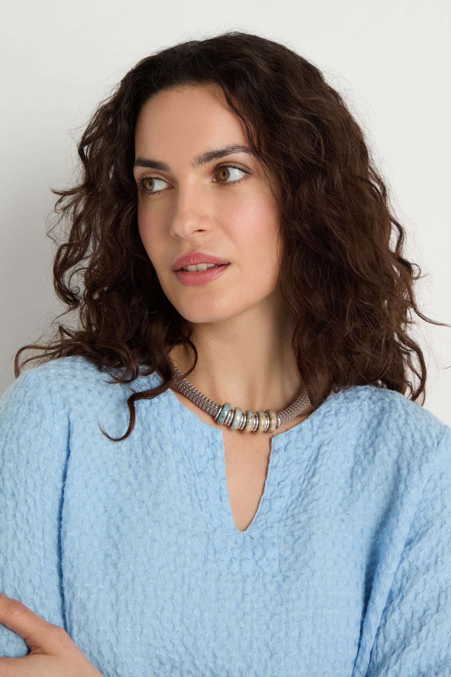 Woman with wavy brown hair wears a textured light blue top and the Mosaic Cylinder Bead Short Cord Necklace, gazing slightly to the side against a plain background.