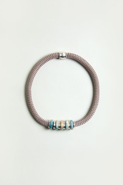 The Mosaic Cylinder Bead Short Cord Necklace features a Sahara exclusive braided taupe cord with a silver clasp and five blue and beige decorative beads, arranged in a circle on a plain light background.