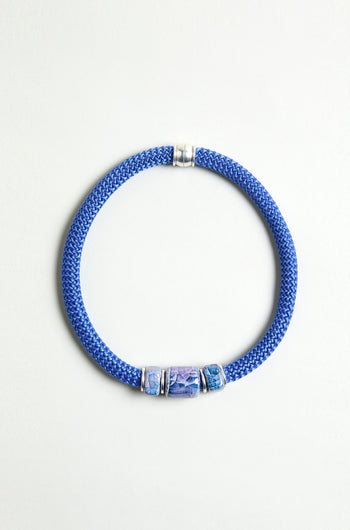 The Triple Marble Bead Short Cord Necklace features three patterned blue and silver cylindrical beads, silver accents, and a chunky cord design, beautifully displayed on a light background.
