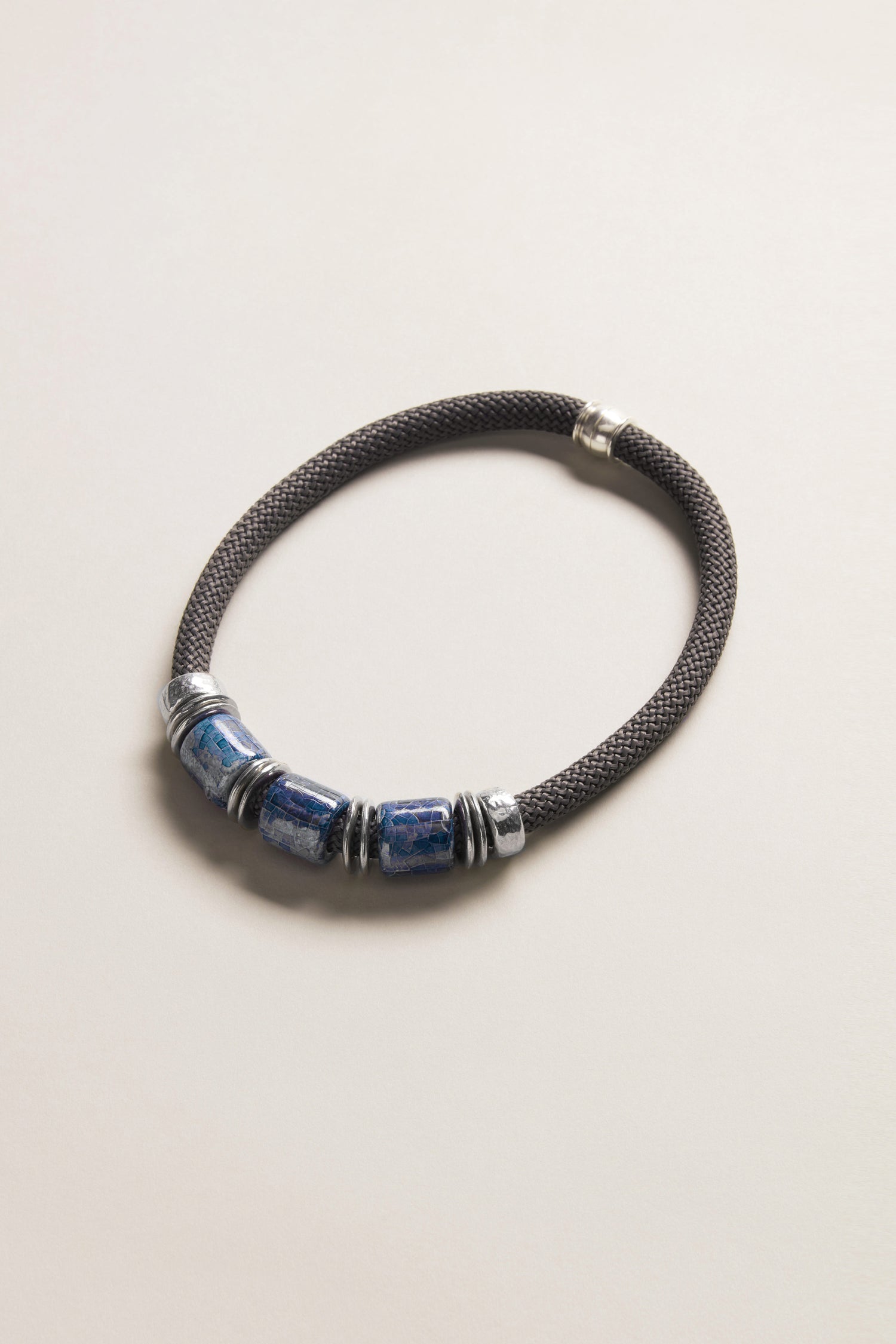 The Trio Mosaic Bead Necklace features three blue ceramic beads and silver-tone metal accents on a dark braided cord, presented against a light neutral background—an elegant example of modern jewelry.