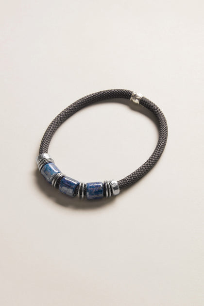The Trio Mosaic Bead Necklace features three blue ceramic beads and silver-tone metal accents on a dark braided cord, presented against a light neutral background—an elegant example of modern jewelry.
