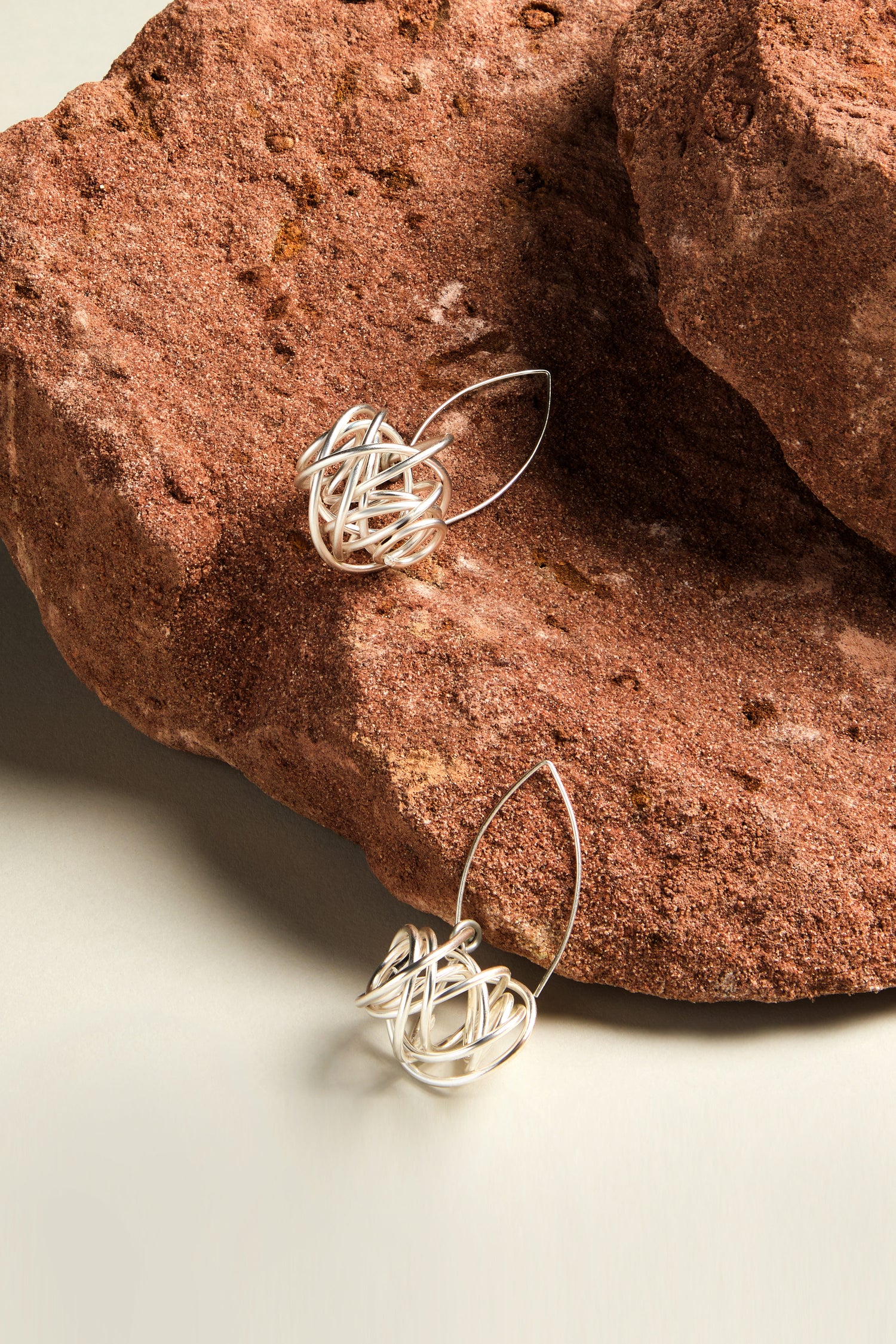 The Silver Scribble Earrings, featuring a knot design, are elegantly showcased on a textured reddish-brown rock surface, forming a modern artistic statement in jewelry.