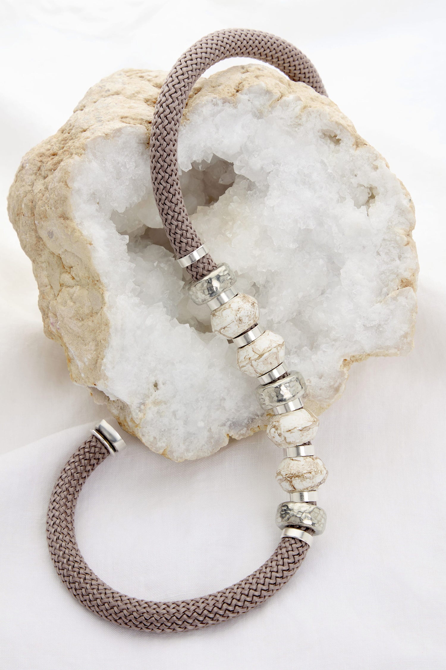 A Marble Bead Cord Necklace with white and silver beads is displayed draped over a textured beige and white rock, reminiscent of its elegant charm.