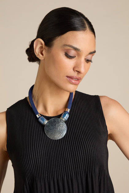 A person with sleek dark hair wears a black pleated top and a blue cord Hammered Sphere Cord Necklace by Etnika Jewellery, featuring a large hammered sphere pendant, looking down slightly.