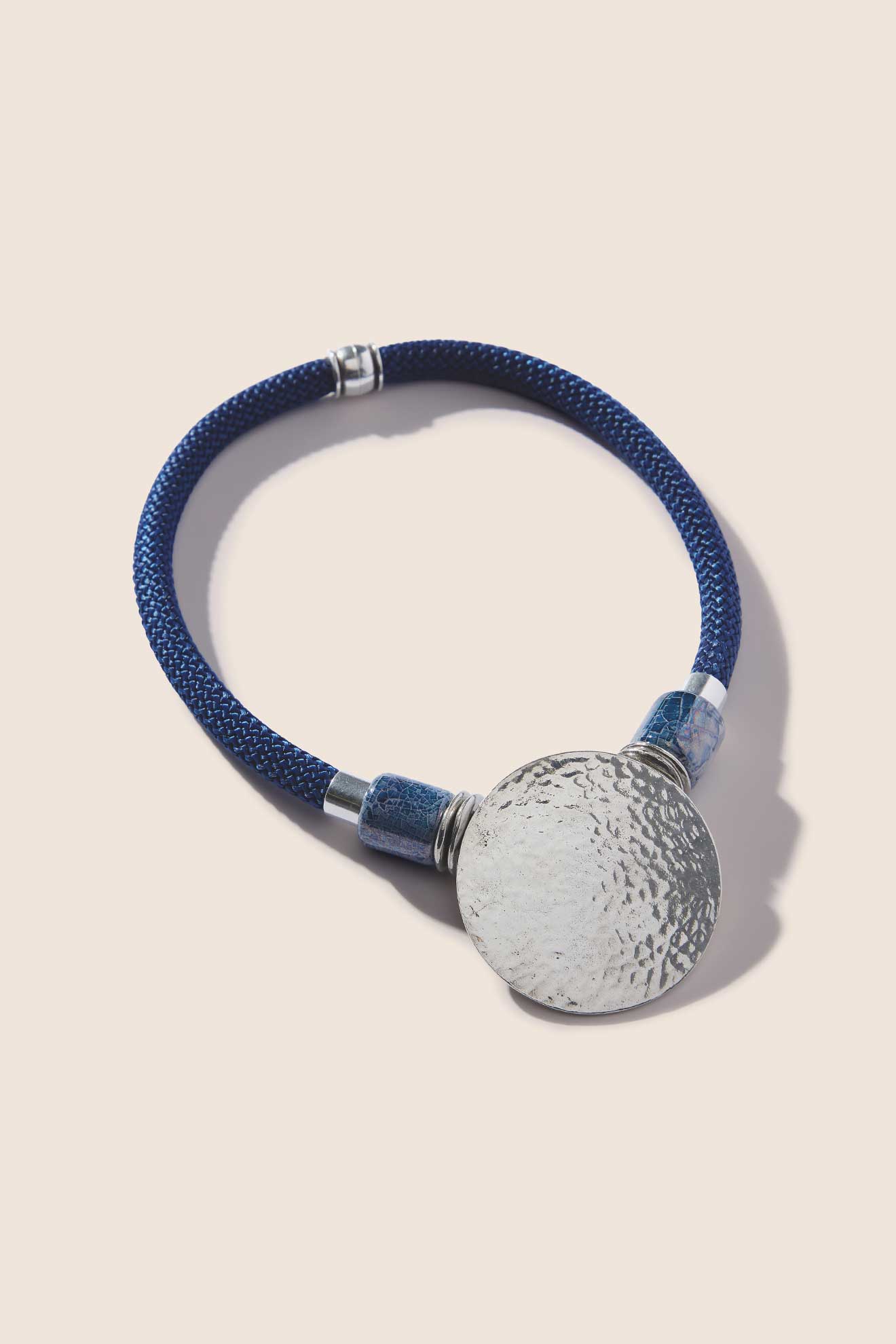 The Hammered Sphere Cord Necklace showcases a round silver pendant with a hammered texture, affixed to a blue braided cord accented with metal details. This statement piece from Etnika jewellery exudes unique charm.