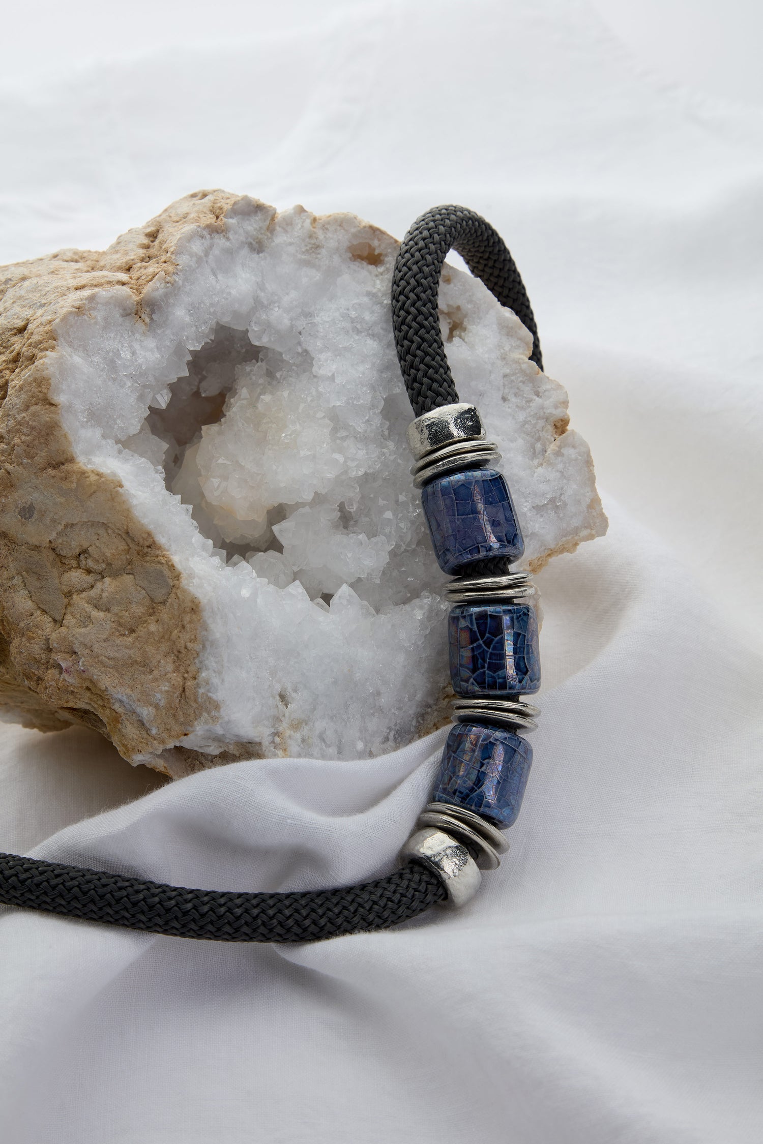 A geode with a hollow center sits on a white fabric, with the Trio Bead Cord Necklace—featuring a black braided cord adorned with three deep blue and silver decorative beads—draped over it, showcasing unique statement art-lead jewellery.