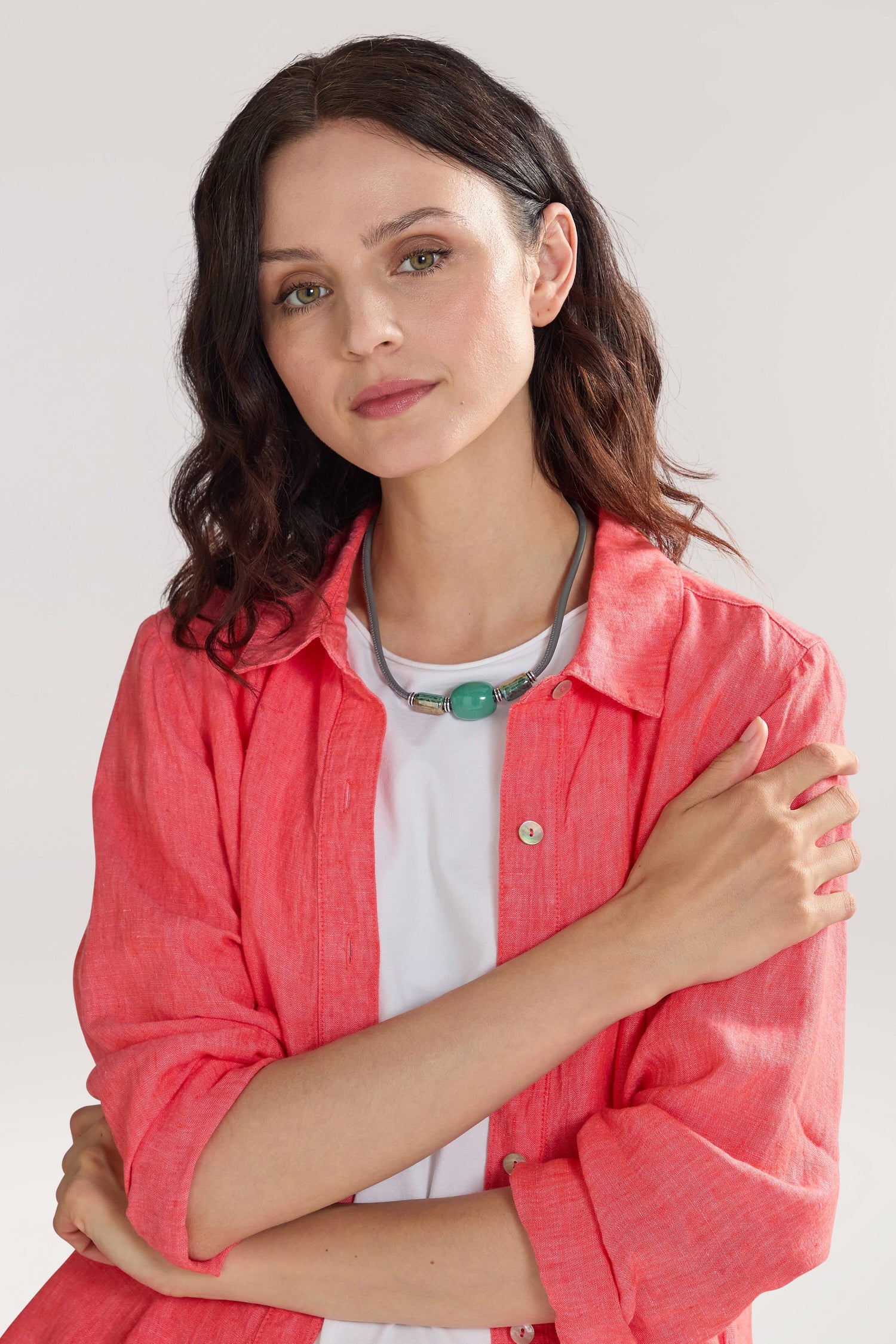 A woman with dark hair wearing a coral-colored shirt over a white top and a Triple Bead Leather Cord Necklace stands with one arm crossed.