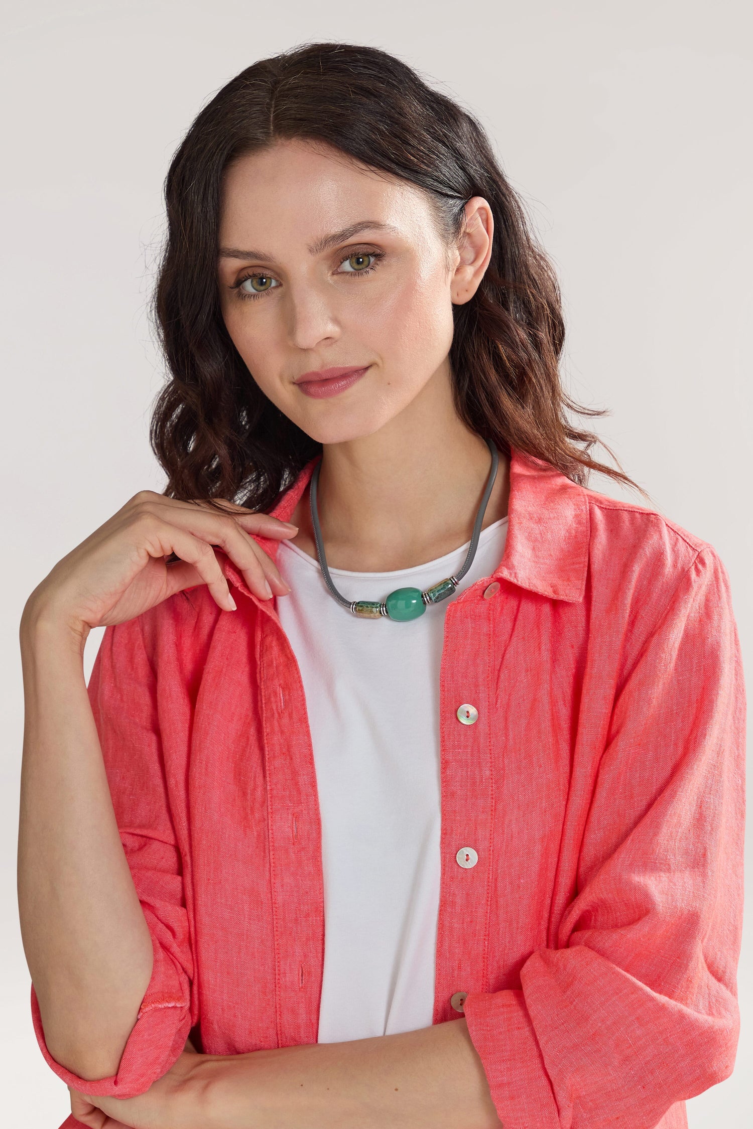 A woman with wavy dark hair wearing a white shirt and a coral pink button-up, complemented by a Triple Bead Leather Cord Necklace featuring blue marbled beads, poses with one hand near her chin against a neutral background.