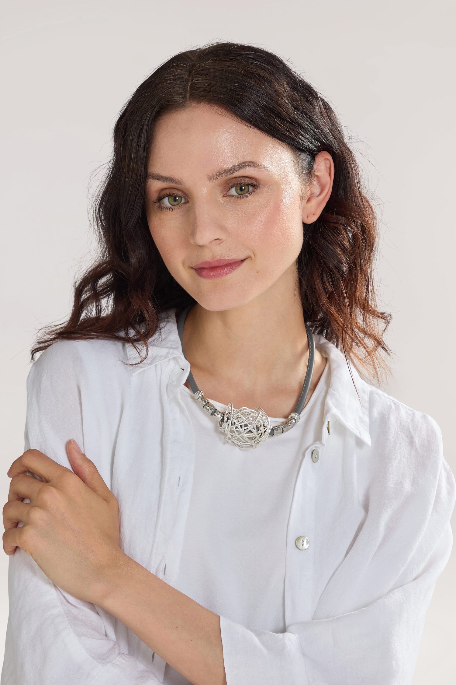 A person with shoulder-length dark hair, wearing a white shirt and a Scribble Bead Leather Cord Necklace, stands with arms crossed against a neutral background.