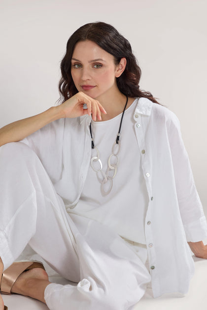 A woman with dark hair, dressed in a white shirt and pants, sits with her elbow resting on her knee and hand near her chin. She is wearing a Silver Links Leather Cord Necklace around her neck as she looks at the camera.