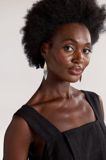 A woman with an afro hairstyle, dressed in a black sleeveless top and Hammered Silver Leaf Earrings, poses against a neutral background.