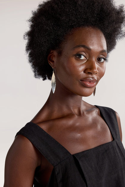 A woman with an afro hairstyle, dressed in a black sleeveless top and Hammered Silver Leaf Earrings, poses against a neutral background.