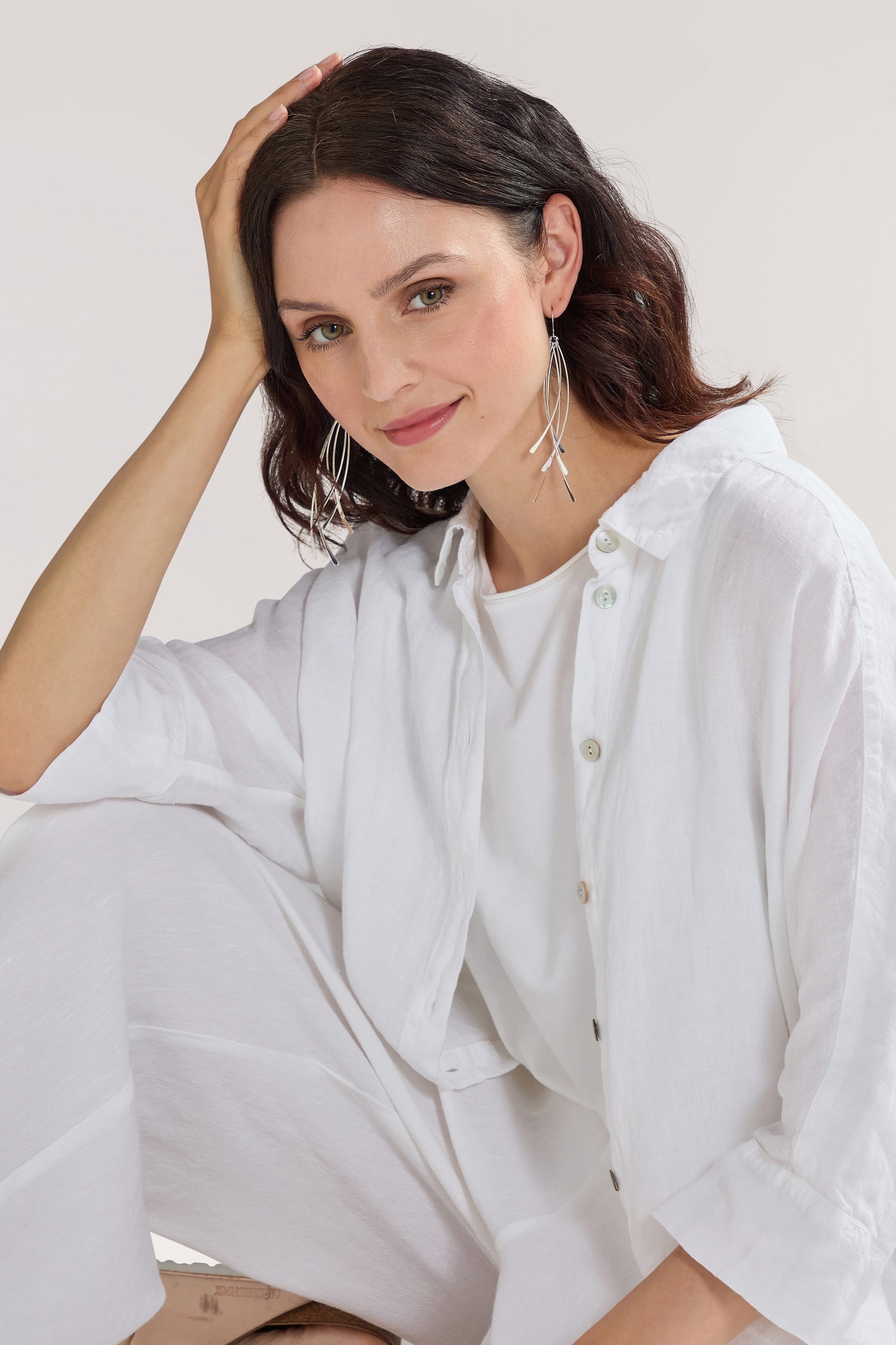 A person with shoulder-length dark hair is sitting with a hand on their head, wearing a white outfit and large hoop Arc Silver Earrings, and smiling slightly.