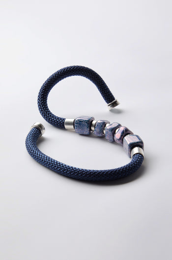 The Mosaic Cube Cord Necklace, featuring a braided blue cord, silver ends, and five marbled beads, displays artisanal craftsmanship on a white background.