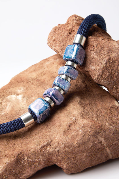 The Mosaic Cube Cord Necklace, featuring navy braided rope and silver-blue patterned beads crafted artisanally, is elegantly draped over a large brown rock.