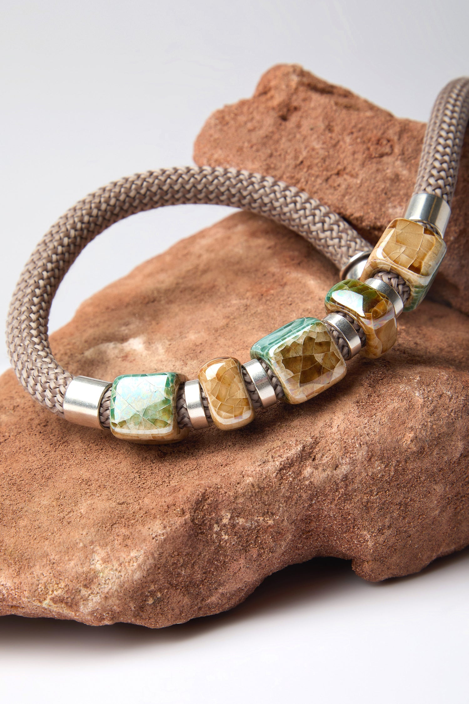 The Marble Cube Cord Necklace is a modern statement piece with square, multi-colored gemstone beads and silver accents, elegantly resting on a textured rock surface. Reflective of Etnika's unique style, it offers a bold and artistic expression.