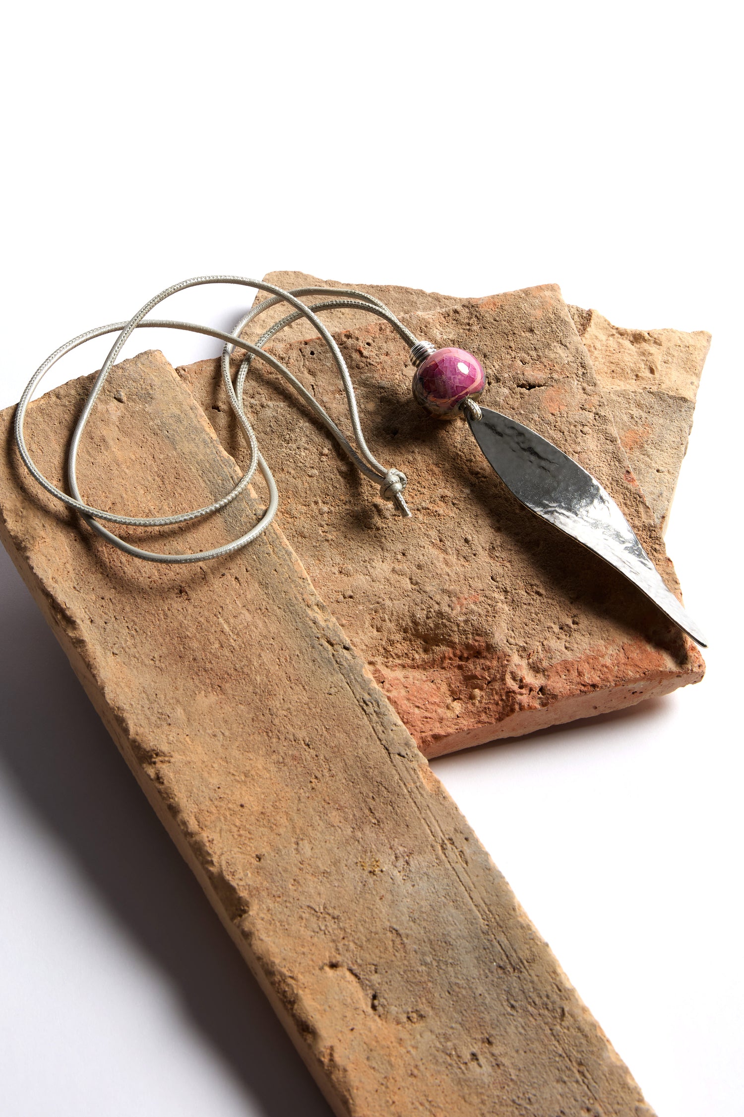 The Twisted Leaf Leather Cord Pendant showcases a silver leaf design with a pink stone on a sleek necklace cord, elegantly set on rough stone slabs, and adds a contemporary touch with its organic shapes.