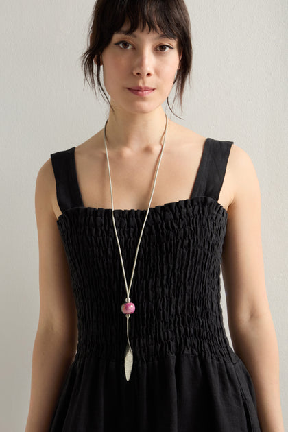 A woman wears a black textured sleeveless dress with a long Etnika Jewelry Twisted Leaf Leather Cord Pendant necklace featuring a pink bead, standing against a plain backdrop.