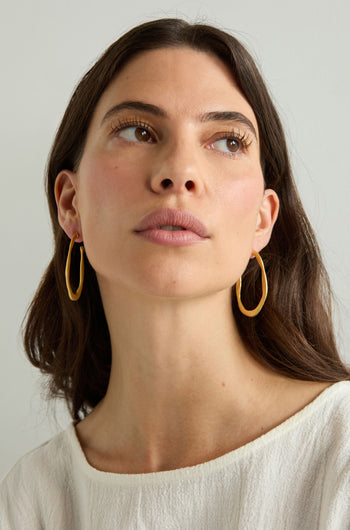 Woman with long brown hair wears Gold Plated Crushed Hoop Earrings and a cream top, gazing upward against a plain light background—a simple showcase of everyday elegance.