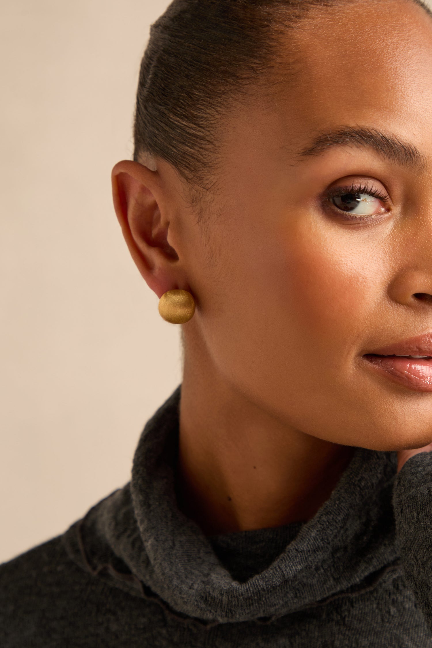 A close-up of a person wearing a gray turtleneck and the Brushed Sphere Earrings, with part of their face, ear, and neck visible against a neutral background. The elegant round gold Brushed Sphere Earrings add a subtle touch of sophistication to the look.