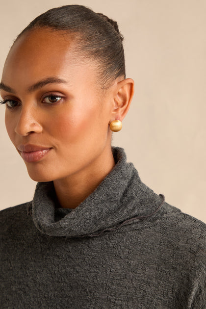 A woman with a neat bun wears a gray turtleneck sweater and the elegant Brushed Sphere Earrings, standing against a plain beige background.