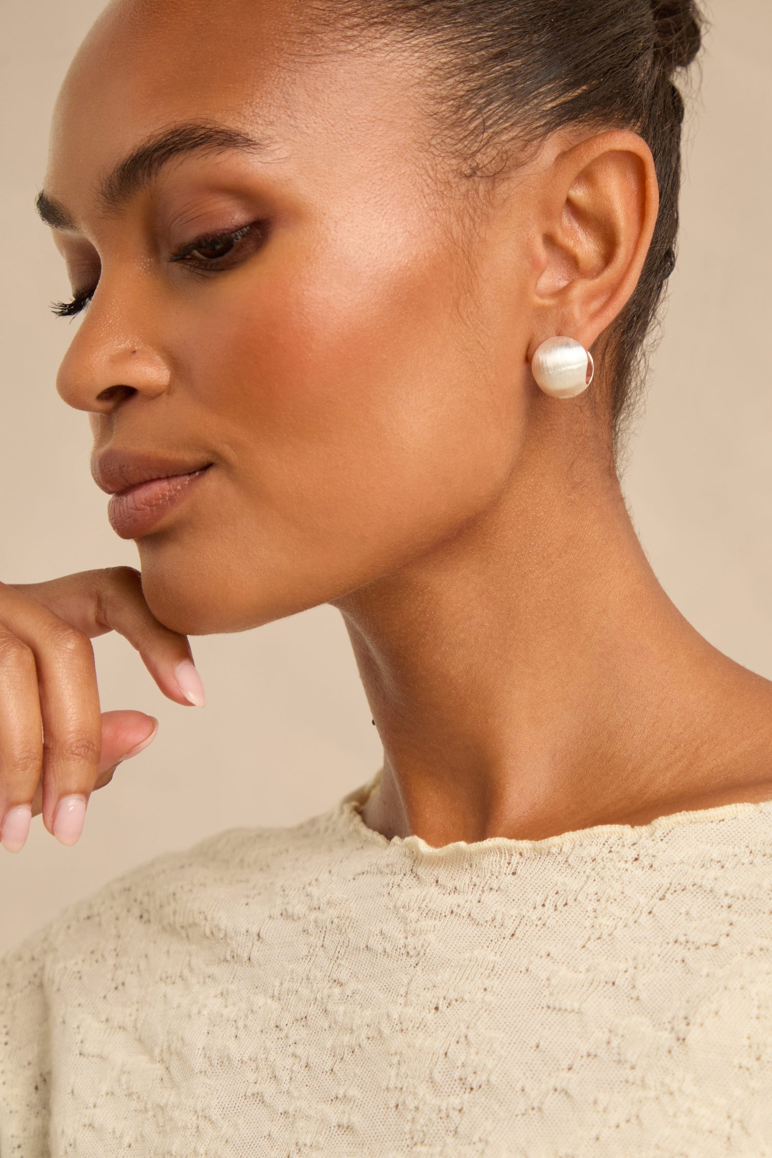 Woman with neat hair and natural makeup, wearing a textured cream top and the Brushed Sphere Earrings—large, round earrings with a brushed finish—rests her chin on her hand.
