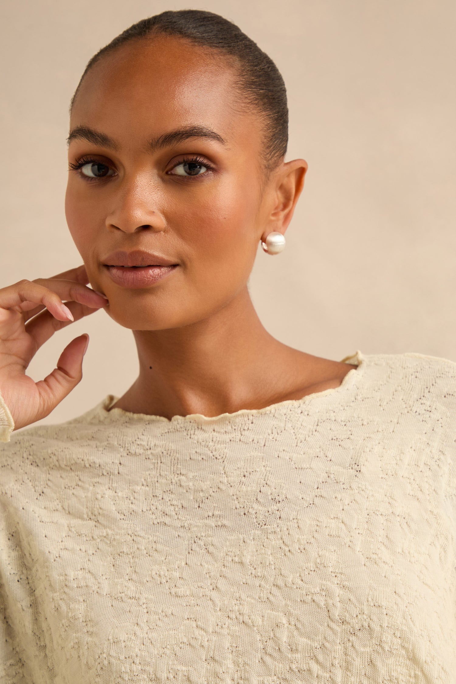 Woman wearing a cream textured top and the Brushed Sphere Earrings, with her hair pulled back, looking at the camera with her hand near her face.