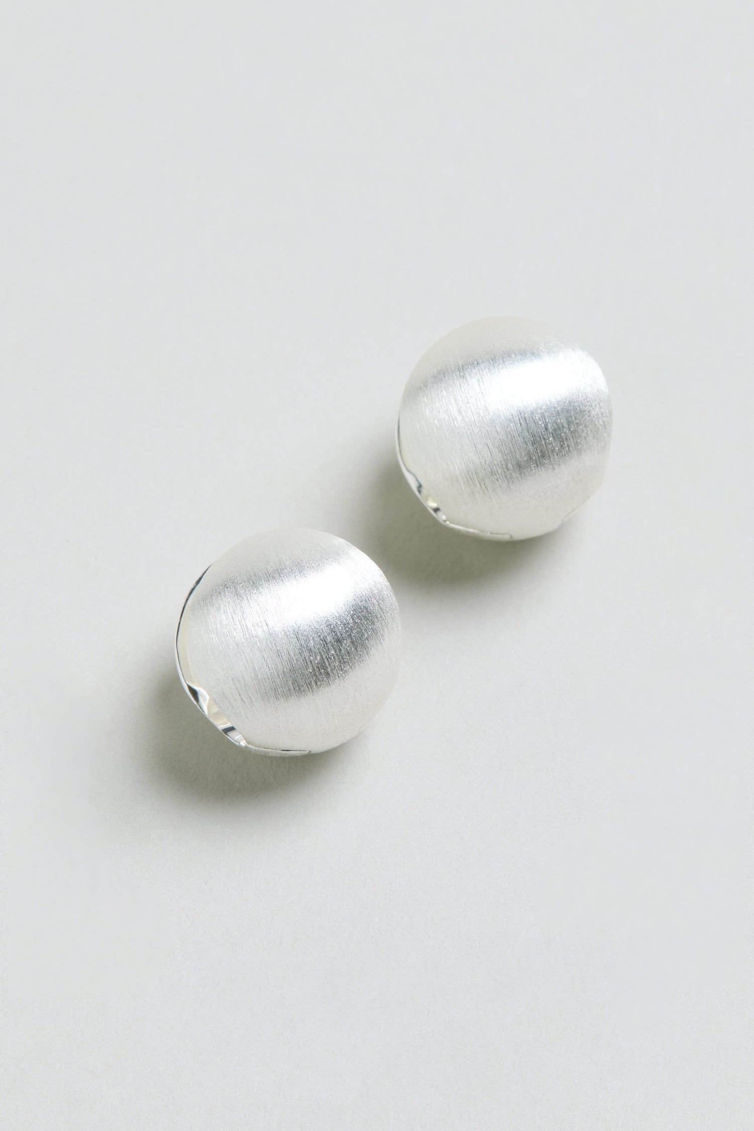 Two Brushed Sphere Earrings, featuring a subtle silver-toned finish and a round, brushed design, are displayed on a light grey background. They are arranged diagonally with a slight gap between them.