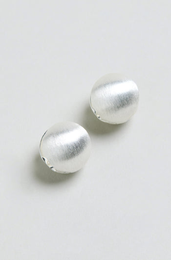 Two Brushed Sphere Earrings, featuring a subtle silver-toned finish and a round, brushed design, are displayed on a light grey background. They are arranged diagonally with a slight gap between them.