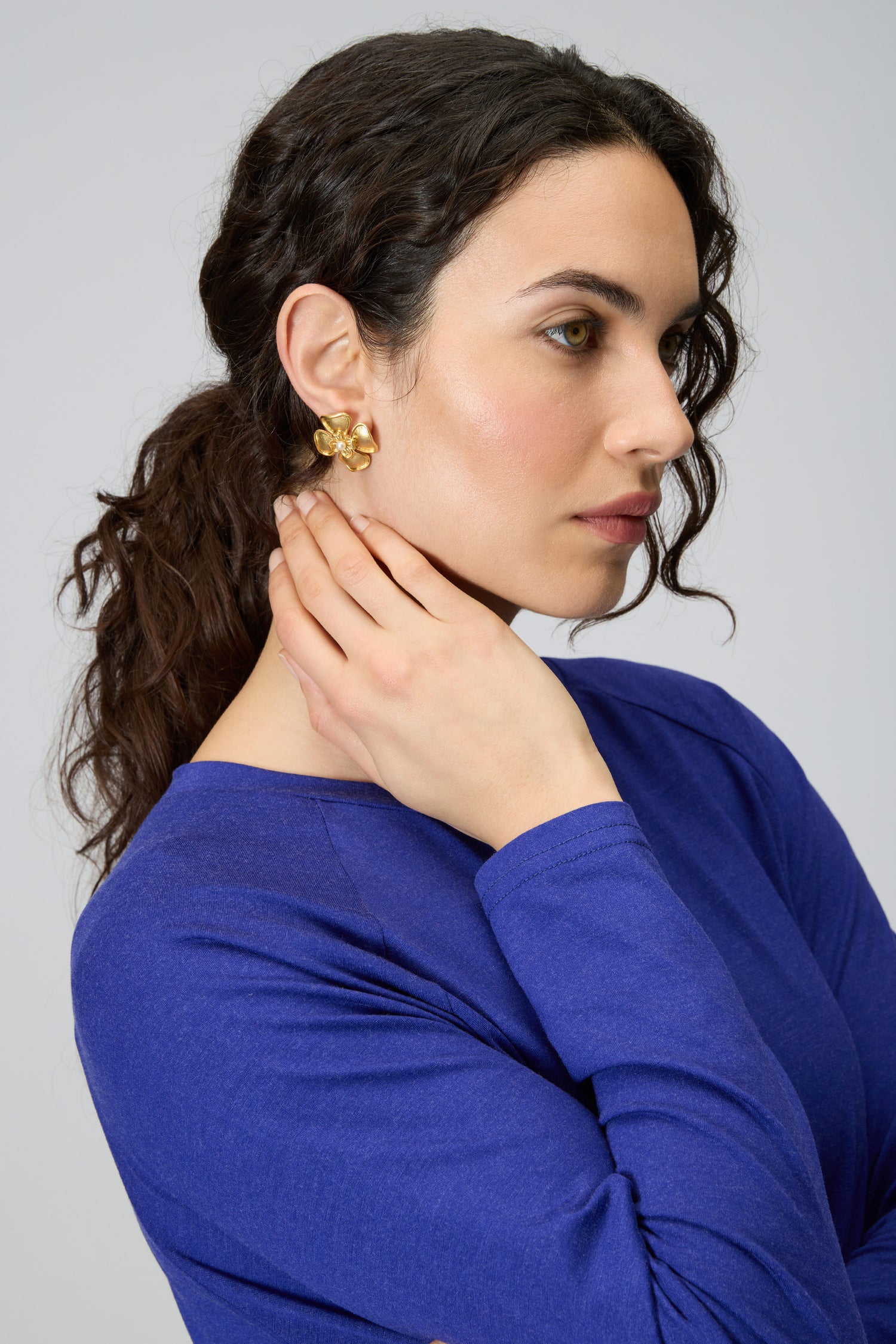 A woman with wavy dark hair, in a blue long-sleeve top and Gold Plated Flower Pearl Earrings, rests her hand on her neck as she gazes to the side.