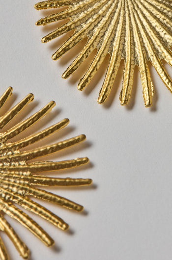 Two textured gold plated Stargazer Earrings, each shaped like a sunburst with radiant lines, rest on a plain light background. Both earrings are partially in frame.