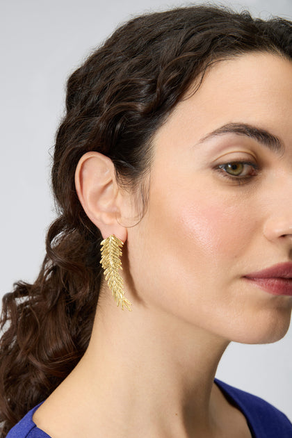 Close-up of a woman with curly brown hair wearing Gold Plated Feather Earrings and a blue top, set against a plain light background.
