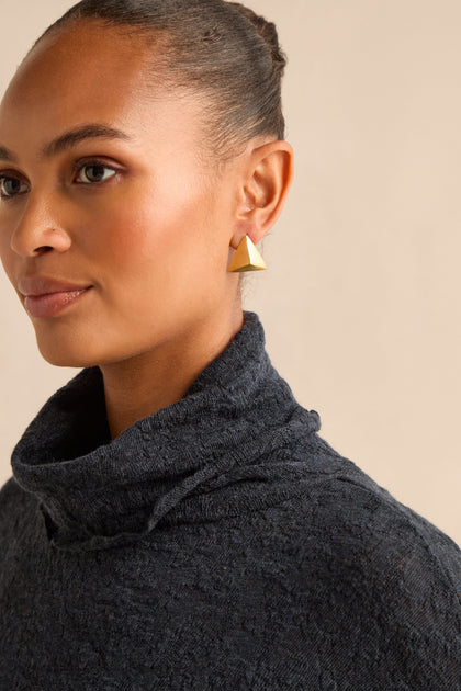 A close-up portrait of a woman with her hair pulled back, wearing a dark grey turtleneck sweater and a single matte gold Folded Triangle Earring.