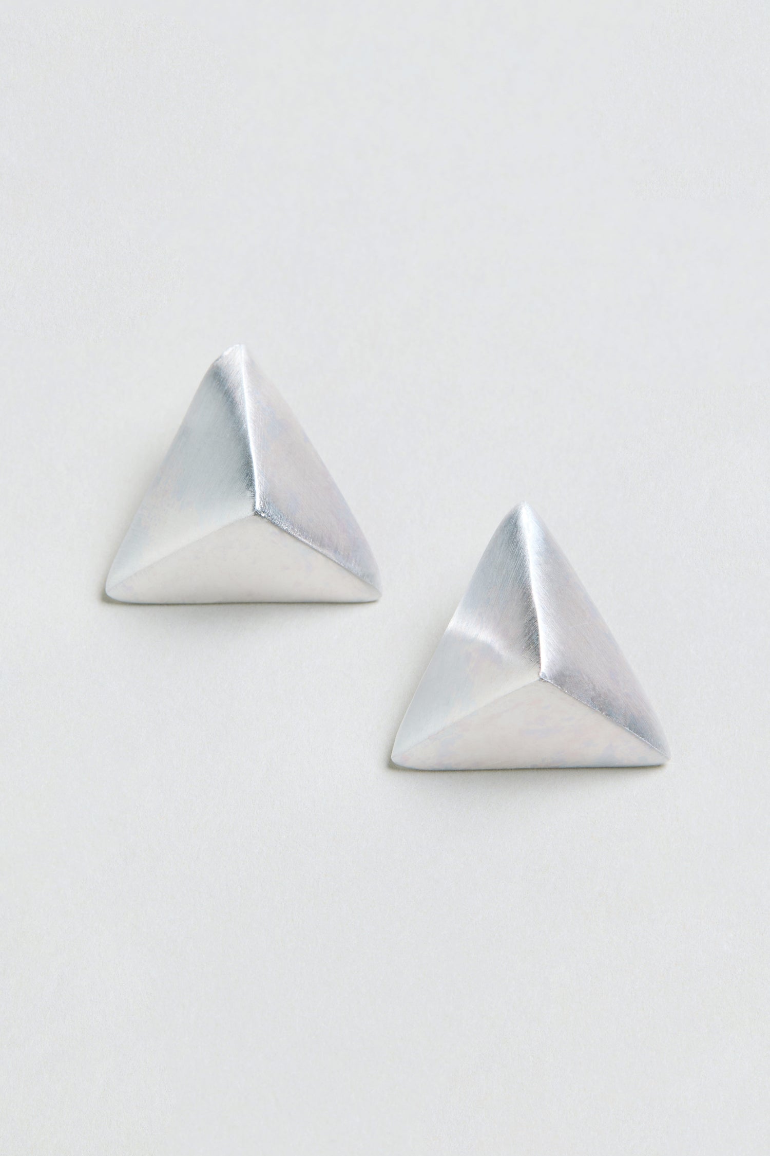 Two silver Folded Triangle Earrings, each featuring a pyramid-shaped geometric design, are displayed against a plain, off-white background.