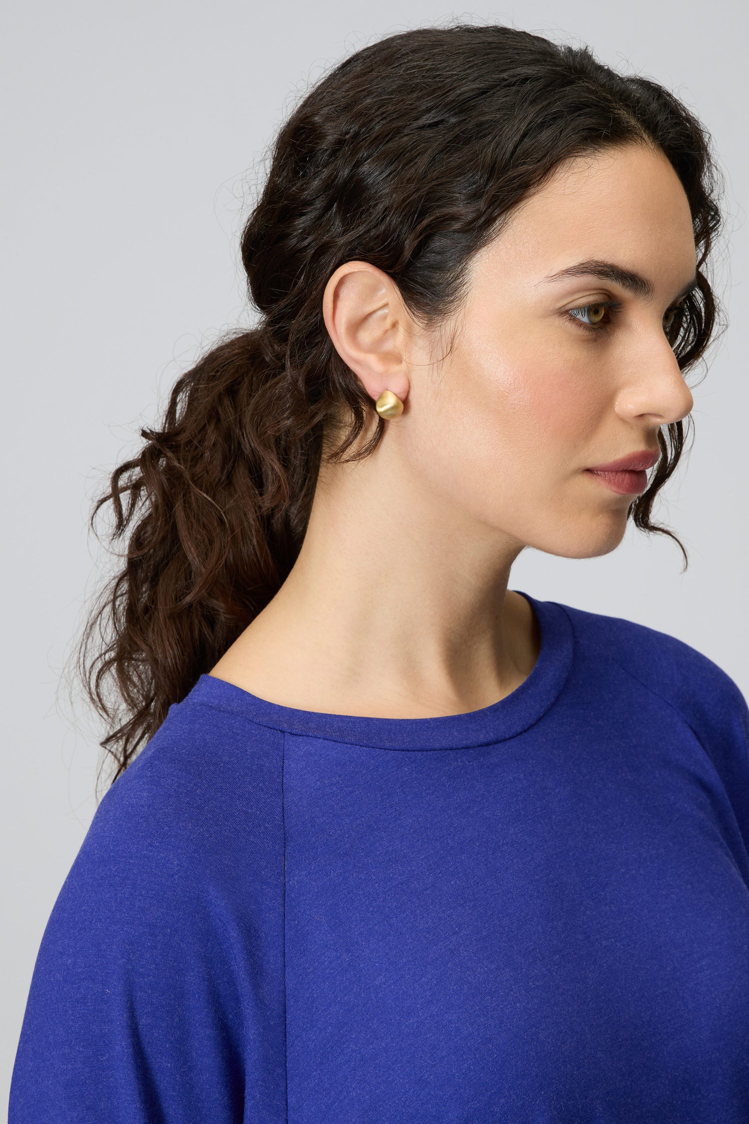 A woman with wavy dark hair in a low ponytail, wearing a blue top and Gold Plated Mini Pebble Earrings, shown in profile against a plain background.
