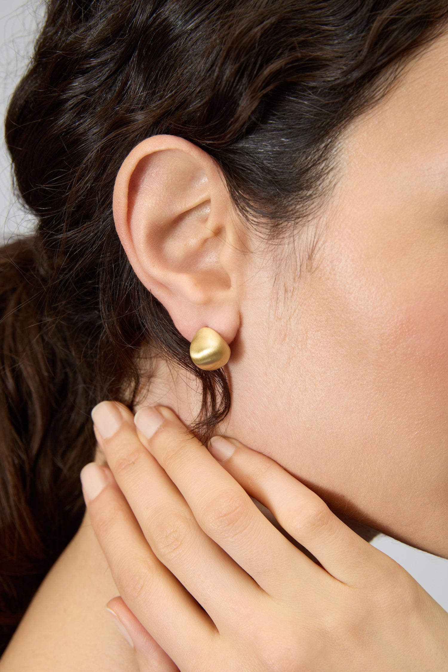 A close-up of a woman's ear wearing Gold Plated Mini Pebble Earrings, her hand gently resting on her neck.