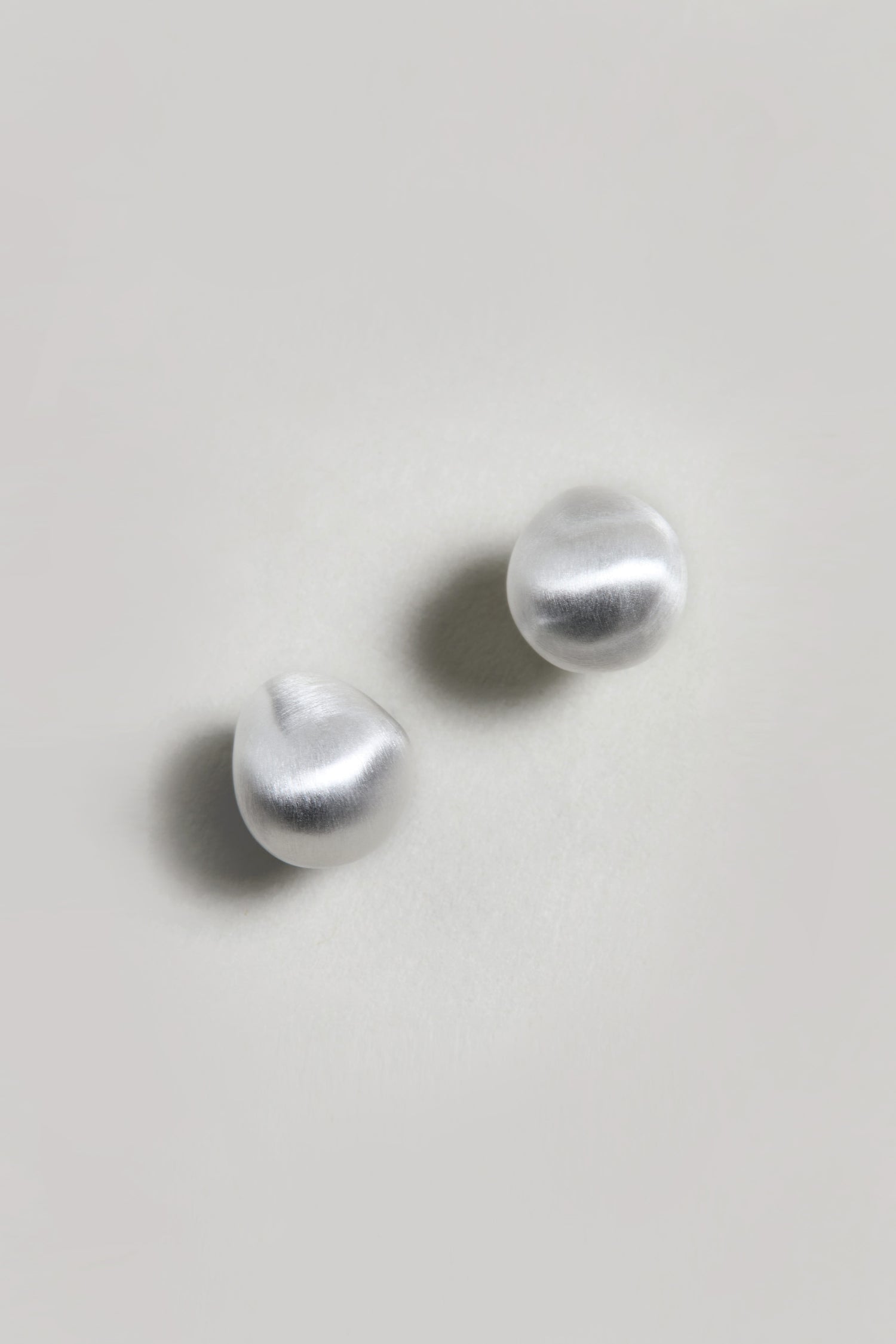The Mini Silver Pebble Earrings, featuring a round, irregular organic shape and a silver-tone finish, are displayed on a plain light-colored surface.