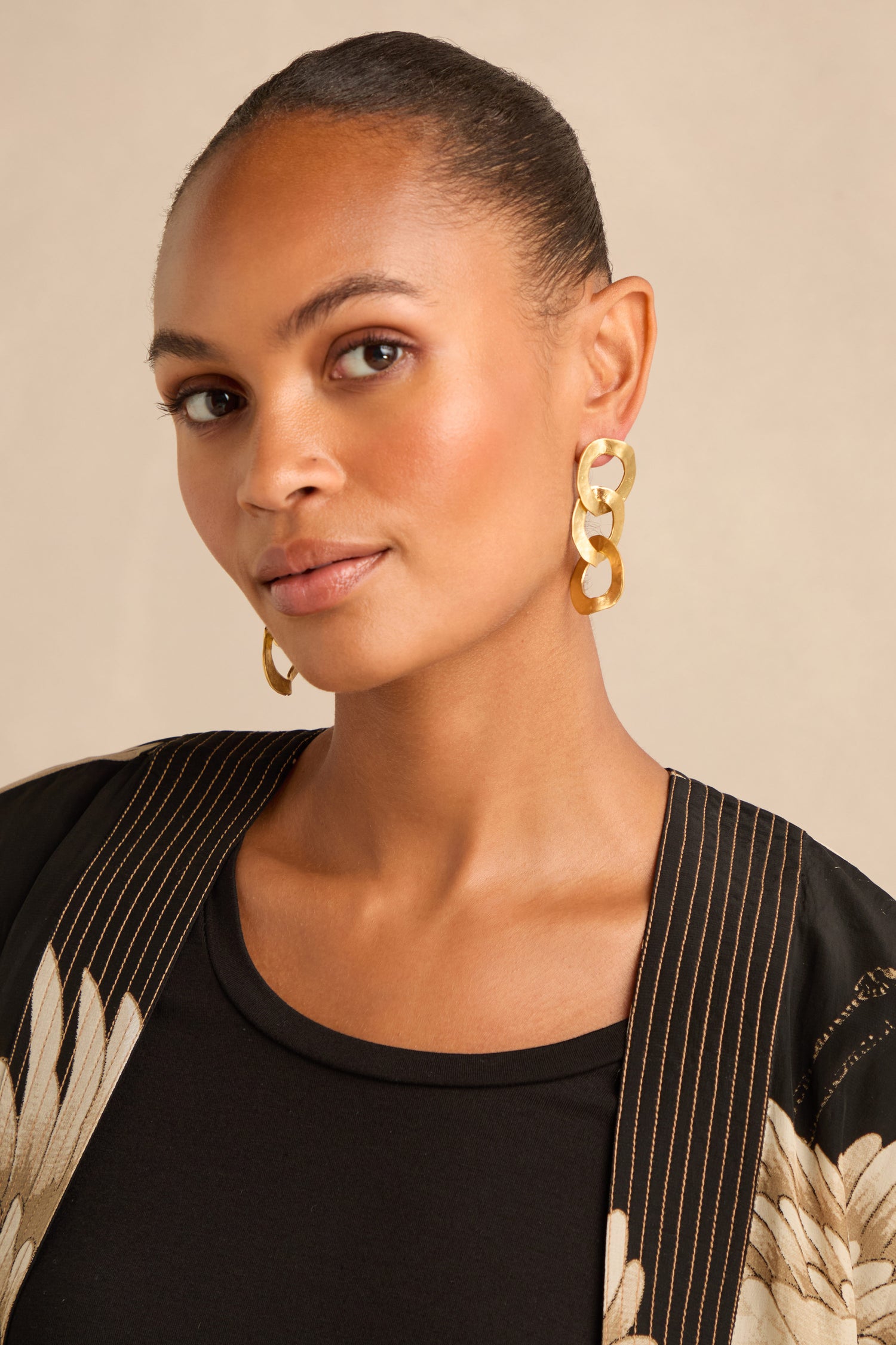 Woman with pulled-back hair wearing Gold Triple Hoop Earrings, a black top, and a patterned black jacket, posing against a neutral background.