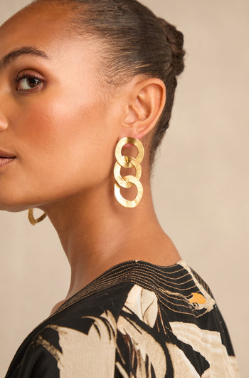A woman with braided hair is shown in profile against a neutral background, wearing Gold Triple Hoop Earrings and a patterned black and beige top.