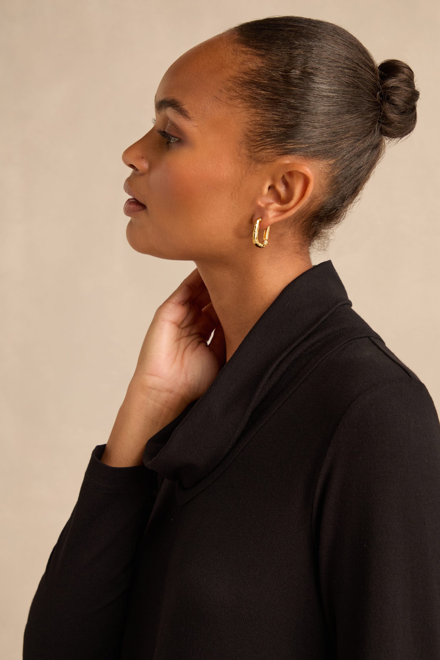 A woman in profile with her hair in a bun wears a black top and the Hammered Hoop Earrings—gold-plated hoops with a hammered texture—as she touches her neck against a neutral background.